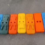 A close-up view of seven colorful plastic fence bases, including blue, green, orange, and yellow, laid out side-by-side on a grey paved surface.