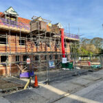 Residential construction site with temporary mesh fencing, scaffolding-covered brick building, red waste chute, and construction notice signs on clear day