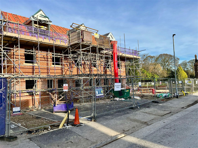 Residential construction site with temporary mesh fencing, scaffolding-covered brick building, red waste chute, and construction notice signs on clear day