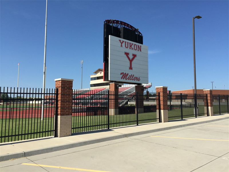 Black steel picket fence with brick pillars securing the perimeter of a large sports stadium or school facility, with a "Yukon Millers" sign and bleachers visible.