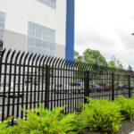 A black metal security fence with curved, pointed tops runs along the perimeter of a building with large windows. Green shrubs line the base of the fence, and a parking lot with cars is visible behind it.
