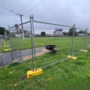 Temporary mesh fencing with yellow concrete bases enclosing a grassy plot of land, with a wheelbarrow inside the fenced area. A sign on the fence displays "0800 TEMPFENCE" and a phone number.