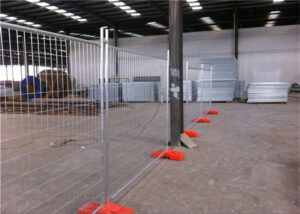 A temporary mesh fence with orange plastic bases set up inside a large industrial warehouse or factory space. A tall metal support pillar stands within the fenced area, and various industrial materials are visible in the background.