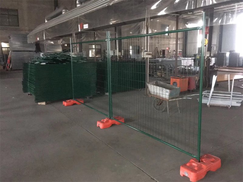 Green powder-coated temporary mesh fencing with orange plastic bases, set up inside a warehouse with stacks of similar green fence panels in the background.