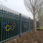 A black metal fence, backed by a green screen, features colorful Olympic rings attached at regular intervals. The fence runs alongside a road with bare trees and dry ground cover, suggesting a public or event-related area.
