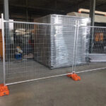 Indoor temporary fencing system with orange bases securing warehouse inventory of wrapped metal panels in industrial facility.