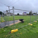 Temporary mesh fencing with yellow plastic bases enclosing a grassy field, with a black wheelbarrow inside and an "ECOTEMP FENCE" sign attached. Houses and power lines are visible in the background.