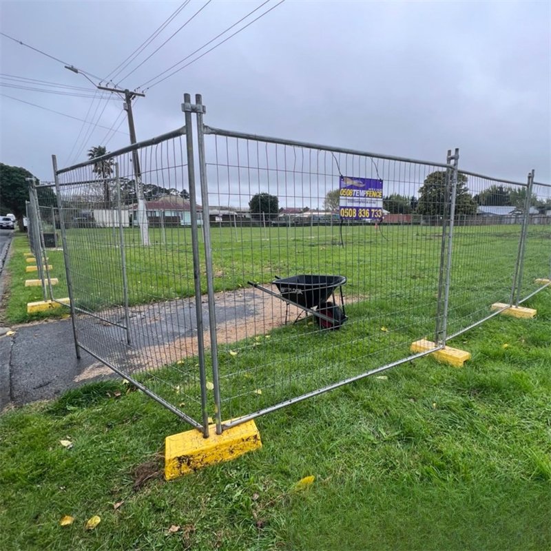 Temporary mesh fencing with yellow plastic bases enclosing a grassy field, with a black wheelbarrow inside and an "ECOTEMP FENCE" sign attached. Houses and power lines are visible in the background.