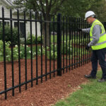 A worker wearing a hard hat and high-visibility vest inspecting the quality and installation of a black steel picket fence in a landscaped area.