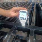 A hand holds a white "Smart Sensor" device, displaying "158" on its screen, against a black metal fence component, likely measuring the coating thickness during manufacturing or quality control.