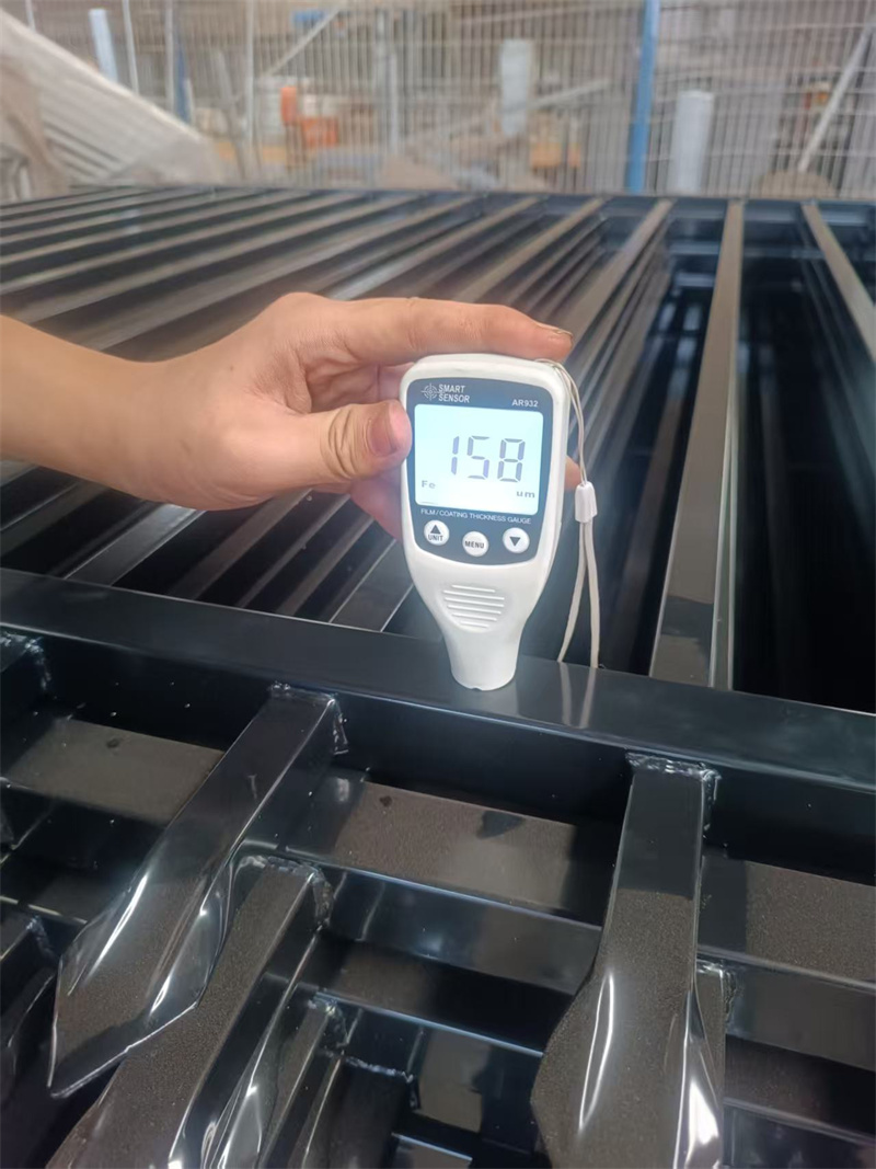 A hand holds a white "Smart Sensor" device, displaying "158" on its screen, against a black metal fence component, likely measuring the coating thickness during manufacturing or quality control.