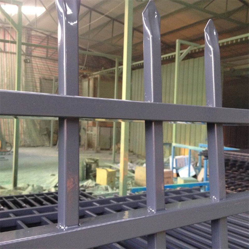 A close-up view of a gray metal fence panel, showing its vertical pickets with sharp, pointed tops and horizontal rails. The background is a blurry industrial setting, likely a manufacturing facility.