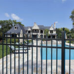 A black metal fence in the foreground provides a view of a luxurious residential backyard. Behind the fence is a sparkling blue swimming pool with a paved deck, lounge chairs, umbrellas, and a large, elegant house surrounded by green trees under a bright blue sky.