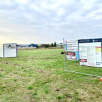 An early-stage construction site with a wide grassy field, featuring two portable site offices and a large temporary safety and information sign framed by temporary fencing.