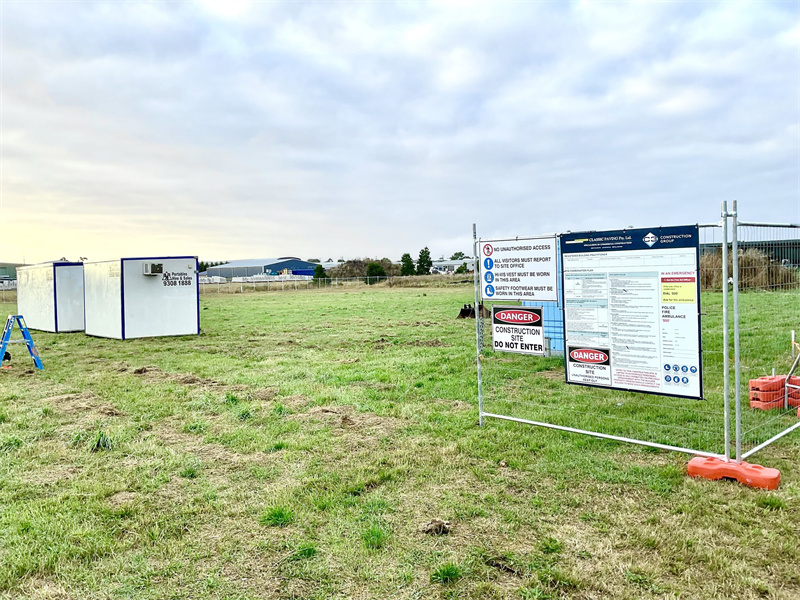 An early-stage construction site with a wide grassy field, featuring two portable site offices and a large temporary safety and information sign framed by temporary fencing.
