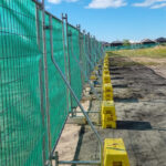 A long line of temporary metal mesh fencing covered with green privacy or shade cloth. The fence is heavily braced with diagonal metal supports and bright yellow plastic bases, some of which are stacked for added stability. The scene is an outdoor construction or development site with dry ground and distant houses under a blue sky with white clouds.