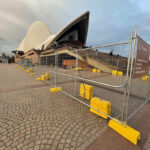 Iconic Sydney Opera House with distinctive white shell architecture surrounded by galvanized steel temporary fencing panels mounted on bright yellow plastic feet, creating restricted access area around entrance with directional signage for Stage Door, Box Office and Bennelong Restaurant visible on cobblestone forecourt under overcast sky.