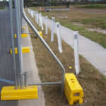 Temporary fencing secured with yellow plastic bases and angled braces, running alongside a paved path and grassy verge with white bollards.
