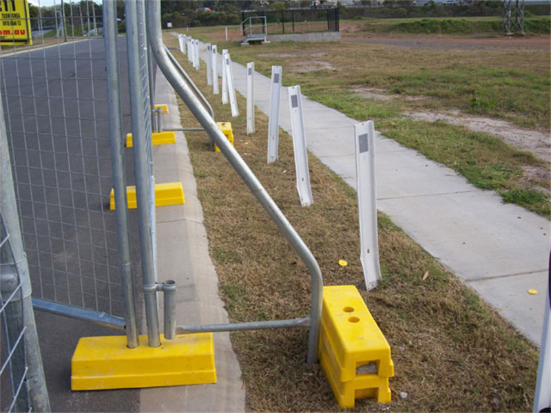 Temporary fencing secured with yellow plastic bases and angled braces, running alongside a paved path and grassy verge with white bollards.