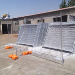 Galvanized temporary fencing panels, some stacked and some erected with orange plastic bases, in an outdoor industrial yard.