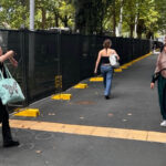 A sidewalk scene with several people walking alongside a long black temporary fence. The fence is supported by bright yellow plastic bases and separates the pedestrian area from a treed background.