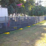 Temporary fencing with black screening mesh and yellow plastic feet enclosing an area next to a white truck, on a grassy field with trees in the background, suggesting an outdoor event or temporary setup.