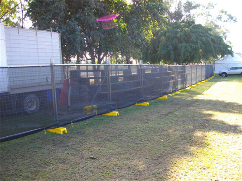 Temporary fencing with black screening mesh and yellow plastic feet enclosing an area next to a white truck, on a grassy field with trees in the background, suggesting an outdoor event or temporary setup.