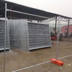A storage area or factory floor showing galvanized temporary fence panels with orange plastic bases, some stacked neatly in the background and one panel assembled in the foreground.