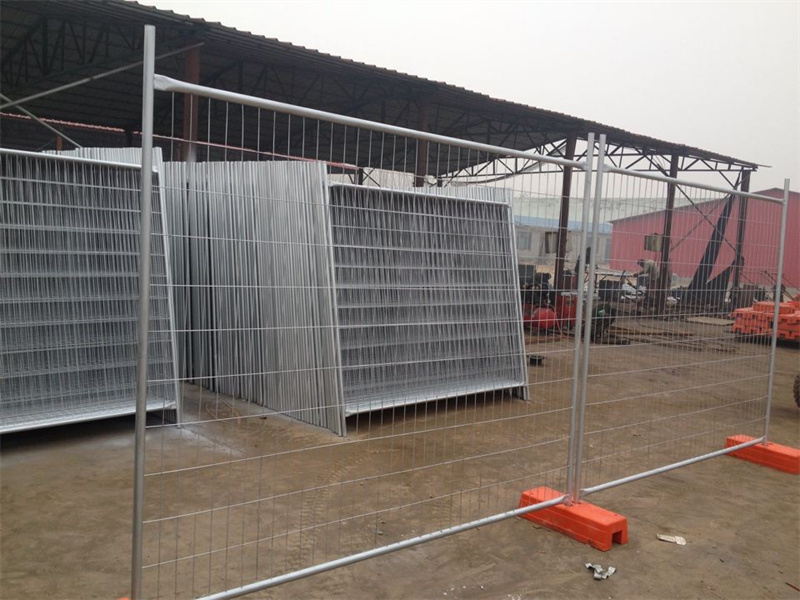 A storage area or factory floor showing galvanized temporary fence panels with orange plastic bases, some stacked neatly in the background and one panel assembled in the foreground.