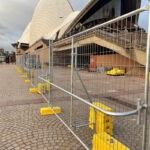 Galvanized temporary fencing with yellow plastic feet forms a barrier in the foreground, with the iconic Sydney Opera House and its distinctive white shell-like roofs visible in the background on a cloudy day.