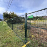 Green temporary mesh fence panels with yellow plastic bases, featuring a "Sunbelt Rentals" logo, installed along a roadside next to an undeveloped grassy area with trees.