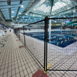 Black temporary fencing with red bases secures a section of a large indoor swimming pool facility, likely for maintenance or an event, with natural light filtering from the roof.
