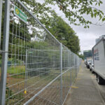 A long line of galvanized temporary mesh fencing with yellow bases extending along a sidewalk, separating a grassy area with trees from a street where a white delivery truck is parked.