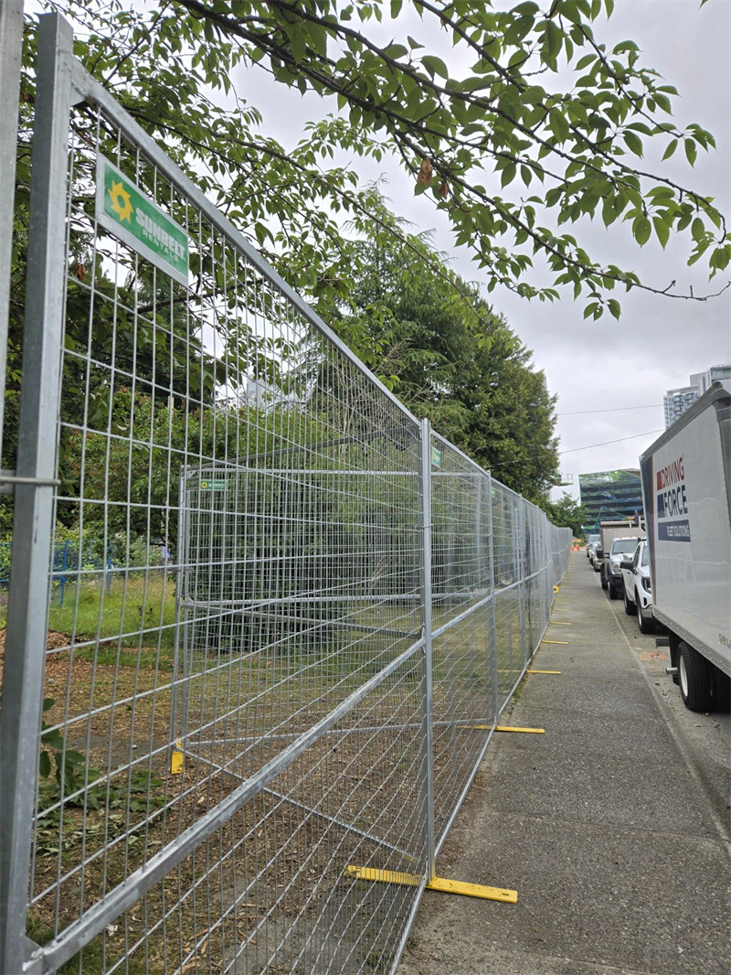 A long line of galvanized temporary mesh fencing with yellow bases extending along a sidewalk, separating a grassy area with trees from a street where a white delivery truck is parked.