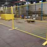 Two yellow temporary welded mesh fence panels with flat metal bases, set up on a factory floor, with more stacked yellow panels in the background.