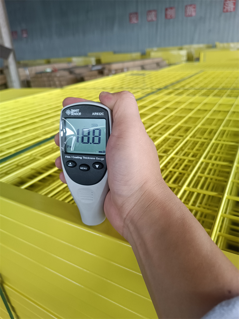 A hand holding a digital coating thickness gauge, displaying '18.8 mil', against a yellow temporary fence panel to measure its surface coating.