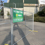 A galvanized temporary fence panel with yellow bases and a green "Sunbelt Rentals - We Rent Fence" sign, placed in front of a building with a green roof, possibly a stadium.