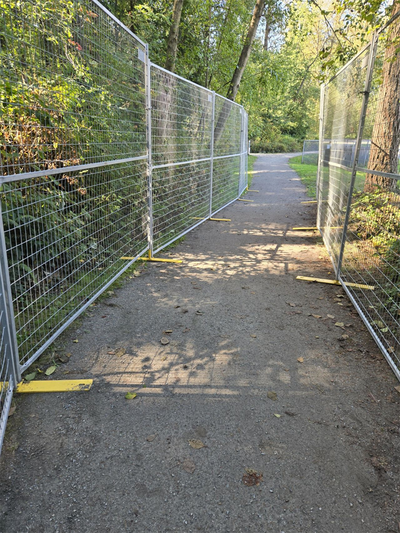 A series of temporary fence panels, light gray in color, line both sides of a gravel pathway, creating a defined walkway through a wooded area with dense green foliage.