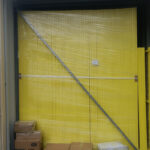 A shipping container is packed with tall stacks of yellow temporary fence panels, individually wrapped in clear plastic, alongside several cardboard boxes.