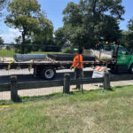 Two construction workers, wearing high-visibility vests, are handling dark temporary fence panels being transported on the back of a flatbed truck. The scene is outdoors near a paved road and a grassy area, with a "SIDEWALK CLOSED" sign visible, suggesting site preparation or removal.