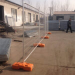 Two panels of temporary fencing with orange plastic feet stand on a concrete ground in an industrial yard, with factory buildings, stacks of metal grates, and a person walking in the background.