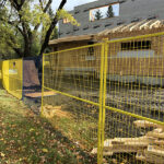 Yellow temporary mesh fencing with bases installed around a residential construction site, where a house frame is being built, with a grassy area and trees visible.