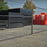 Black temporary fence panels with yellow bases, featuring a "Sunbelt Rentals" logo, separate a gravel area from stacks of construction materials and orange barriers in the background.
