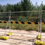 Galvanized steel mesh temporary fencing panels mounted on yellow concrete feet with black and yellow diagonal hazard warning tape, installed along paved walkway adjacent to lush green vegetation and mature trees on sunny day.