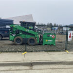 A green temporary fence with yellow bases enclosing a green Kubota skid steer loader and a green Sunbelt Rentals waste bin on a gravel surface. A black van and a building under construction are visible in the background under an overcast sky.