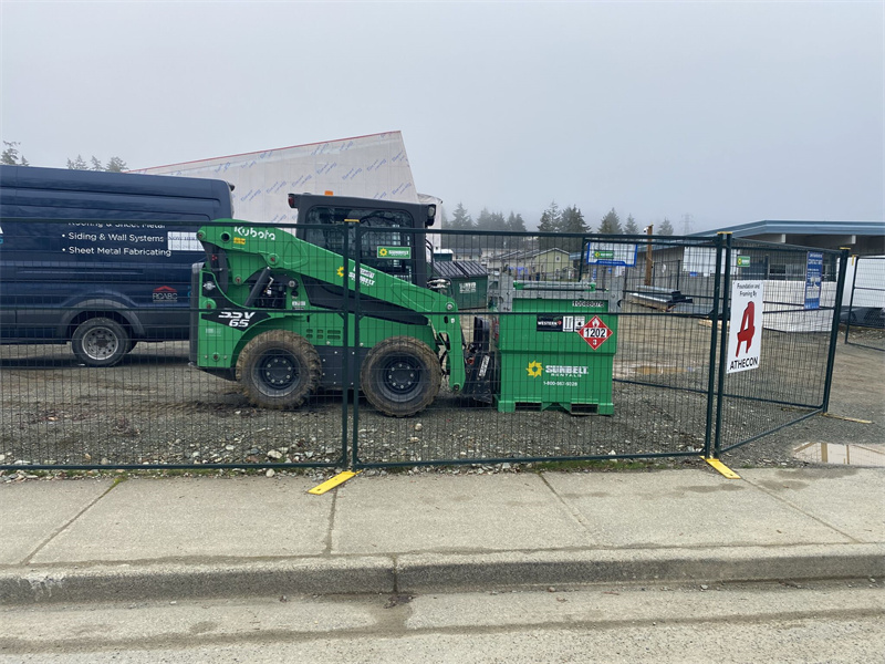 A green temporary fence with yellow bases enclosing a green Kubota skid steer loader and a green Sunbelt Rentals waste bin on a gravel surface. A black van and a building under construction are visible in the background under an overcast sky.