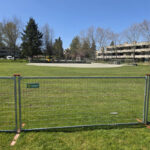 Galvanized temporary welded mesh fence panels with orange connecting clips and bases, set up on a green grassy field under a clear blue sky, with trees and buildings in the background. A "SHOREFLY" logo is visible on the fence.