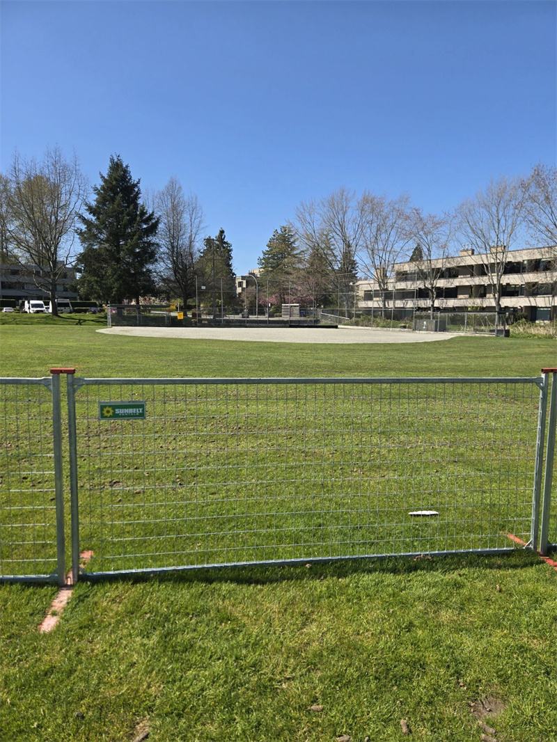 Galvanized temporary welded mesh fence panels with orange connecting clips and bases, set up on a green grassy field under a clear blue sky, with trees and buildings in the background. A "SHOREFLY" logo is visible on the fence.