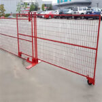Red temporary fencing with welded wire mesh, including a gate section equipped with wheels for easy opening, installed in a paved parking lot with cars visible in the background.