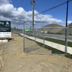 Temporary galvanized wire mesh fencing is installed along the edge of a construction or development site next to a paved road. A "Coming Soon" sign for "Dynamic Commercial Spaces For Lease" stands in the foreground, while piles of earth and gravel are visible in the background under a clear blue sky.